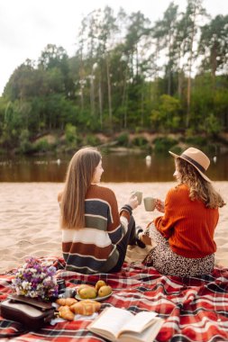 İki genç kadın sahilde piknik yapıyor. Termos ve kahveyle kırmızı hasırın üzerinde oturan pozitif modeller. Dişiler mini piknikte doğanın tadını çıkarırlar. Rahatlama kavramı, doğa..