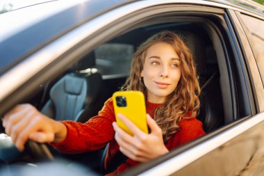 Arabada oturan genç bir kadın sürücü, akıllı telefon kullanıyor. E-posta sohbetlerini kontrol edip, haberleri okuyorum. Seyahat, teknoloji, internet kavramı.