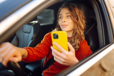 Arabada oturan genç bir kadın sürücü, akıllı telefon kullanıyor. E-posta sohbetlerini kontrol edip, haberleri okuyorum. Seyahat, teknoloji, internet kavramı.
