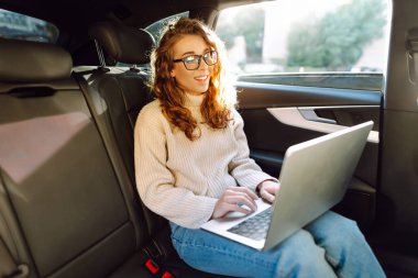 Güzel bir kadın arabanın arka koltuğunda otururken dizüstü bilgisayar kullanıyor. Uzaktan kumanda. Seyahat, teknoloji, internet kavramı.