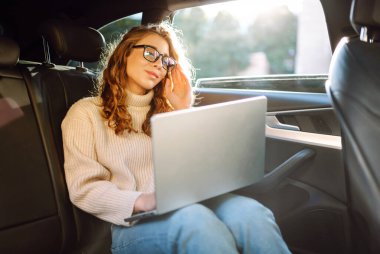 Güzel bir kadın arabanın arka koltuğunda otururken dizüstü bilgisayar kullanıyor. Uzaktan kumanda. Seyahat, teknoloji, internet kavramı.