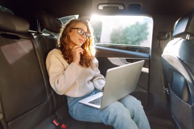 Güzel bir kadın arabanın arka koltuğunda otururken dizüstü bilgisayar kullanıyor. Uzaktan kumanda. Seyahat, teknoloji, internet kavramı.