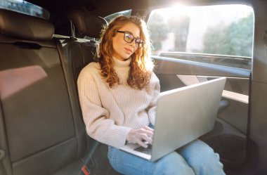 Güzel bir kadın arabanın arka koltuğunda otururken dizüstü bilgisayar kullanıyor. Uzaktan kumanda. Seyahat, teknoloji, internet kavramı.