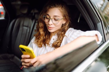 Genç bir kadın seyahat ederken gülümseyip cep telefonu kullanıyor. İş, teknoloji, seyahat, online iletişim kavramı. Taksi..