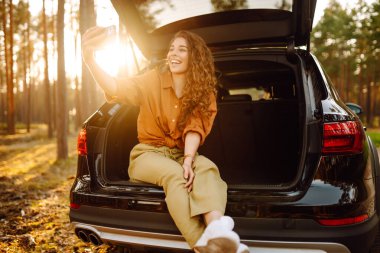 Neşeli kadın gezgin bagajda oturmuş seyahat ederken akıllı telefondan selfie çekiyor. Yaşam tarzı, seyahat, turizm, doğa, aktif yaşam.