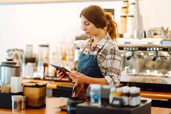 Smiling female barista takes an order from a tablet while standing at the bar counter in a coffee shop. Online order. Small business, people, takeaway and service concept.