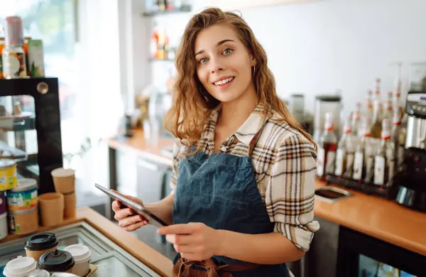 Smiling female barista takes an order from a tablet while standing at the bar counter in a coffee shop. Online order. Small business, people, takeaway and service concept.