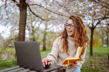 Genç bir kız öğrenci elinde dizüstü bilgisayarla parkta oturuyor. Eğitim çevrimiçi. İş, blog veya serbest çalışma kavramı.