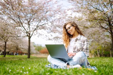 Genç bir kız öğrenci elinde dizüstü bilgisayarla parkta oturuyor. Eğitim çevrimiçi. İş, blog veya serbest çalışma kavramı.