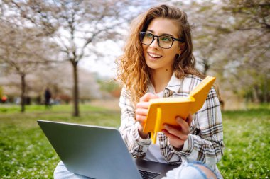 Genç bir kız öğrenci elinde dizüstü bilgisayarla parkta oturuyor. Eğitim çevrimiçi. İş, blog veya serbest çalışma kavramı.