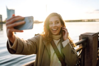 Cep telefonu olan mutlu bir kadın, selfie çekiyor, arkadaşlarıyla iletişim kuruyor, dışarıda blog yazıyor. Yaşam tarzı, seyahat, turizm, doğa, aktif yaşam.