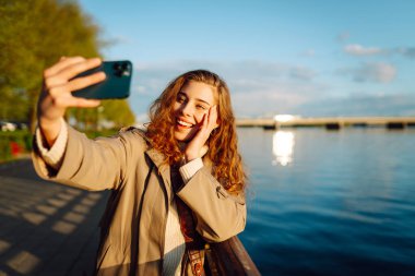 Cep telefonu olan mutlu bir kadın, selfie çekiyor, arkadaşlarıyla iletişim kuruyor, dışarıda blog yazıyor. Yaşam tarzı, seyahat, turizm, doğa, aktif yaşam.