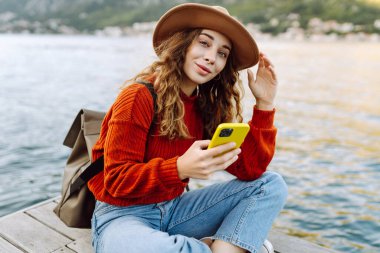 Genç bayan turist okyanus kıyısında akıllı telefon kullanıyor. Yaşam tarzı, seyahat, turizm, aktif yaşam.
