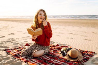 Plaj pikniğinde battaniyeye oturmuş kitap okuyan güzel bir kadın. Rahatlama, keyif, doğayla baş başa kalma kavramı..