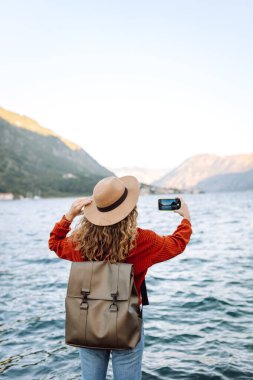 Gündelik süveter giyen kadın gezgin tatilde dağlarla, okyanuslarla ve günbatımında gökyüzüyle muhteşem manzaranın fotoğrafını çekiyor. Tatil sırasında turist..