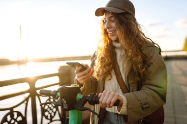 Akıllı telefondan elektrikli scooter çalıştırılıyor. Şehirde elektrikli scooter kullanan mutlu bir kadın. Ekolojik ulaşım konsepti. Seyahat, turizm konsepti. 
