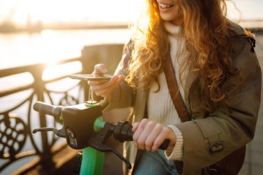 Akıllı telefondan elektrikli scooter çalıştırılıyor. Şehirde elektrikli scooter kullanan mutlu bir kadın. Ekolojik ulaşım konsepti. Seyahat, turizm konsepti. 