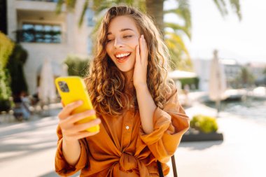 Tatilde gülümseyen genç bir kadın. Yüzünde gülümsemeyle şehrin caddesinde akıllı telefon kullanarak dikiliyorsun. Seyahat, moda, blog konsepti.