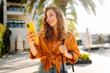 Tatilde gülümseyen genç bir kadın. Yüzünde gülümsemeyle şehrin caddesinde akıllı telefon kullanarak dikiliyorsun. Seyahat, moda, blog konsepti.