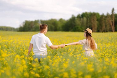 Birbirine aşık genç bir çift kolza tohumu tarlasında yürüyor. Aşk kavramı, ilişki. Moda, güzellik konsepti. Yaz manzarası.