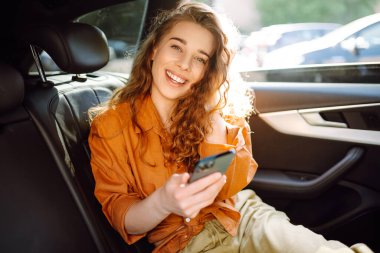 Genç bir kadın arabanın arka koltuğunda otururken akıllı telefon kullanır. Teknoloji kavramı, araba ve iş seyahati..