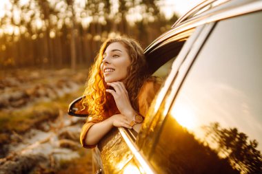 Genç kadın arabada dinleniyor ve gün batımının tadını çıkarıyor. Yaşam tarzı, seyahat, turizm, doğa, aktif yaşam.