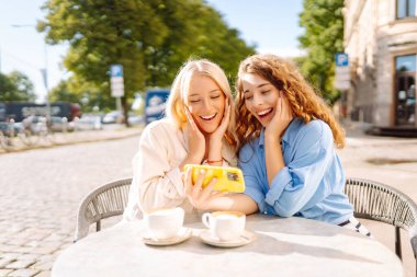 İki gülümseyen kadın bir kafede oturuyor, kahve içiyor, selfie çekiyor, arkadaşlarıyla konuşuyor. Moda, güzellik, blog, turizm.