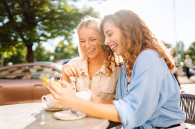 İki gülümseyen kadın bir kafede oturuyor, kahve içiyor, selfie çekiyor, arkadaşlarıyla konuşuyor. Moda, güzellik, blog, turizm.