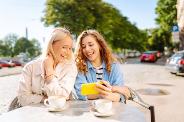 İki gülümseyen kadın bir kafede oturuyor, kahve içiyor, selfie çekiyor, arkadaşlarıyla konuşuyor. Moda, güzellik, blog, turizm.