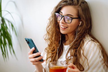 Elinde çay tutan ve akıllı telefon kullanan mutlu bir kadın. İletişim, boş zaman, yaşam tarzı ve ev hayatı.