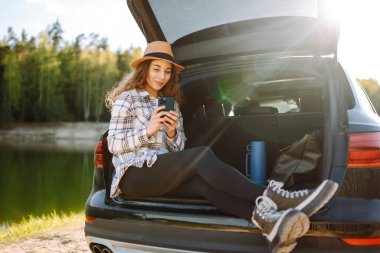 Elinde telefon olan mutlu bir kadın bir arabanın bagajında oturuyor. Teknoloji kavramı, macera, arabayla seyahat..