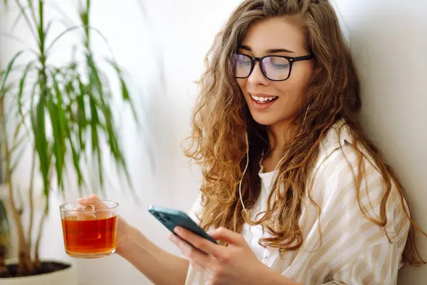 stock image Happy woman holding tea and using smartphone sitting at home. Communication, free time, lifestyle and domestic life.