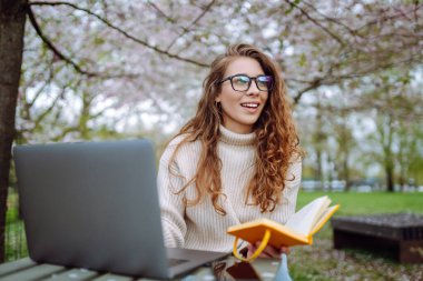 Kampüs parkındaki yeşil çayır ödevini yaparken dizüstü bilgisayar okuyan pozitif bir öğrenci. Eğitim çevrimiçi. IT, uzaktan kumanda. İş, blog yazma, serbest çalışma kavramı.