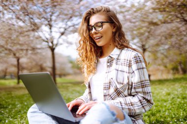 Kampüs parkındaki yeşil çayır ödevini yaparken dizüstü bilgisayar okuyan pozitif bir öğrenci. Eğitim çevrimiçi. IT, uzaktan kumanda. İş, blog yazma, serbest çalışma kavramı.