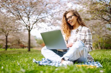 Kampüs parkındaki yeşil çayır ödevini yaparken dizüstü bilgisayar okuyan pozitif bir öğrenci. Eğitim çevrimiçi. IT, uzaktan kumanda. İş, blog yazma, serbest çalışma kavramı.