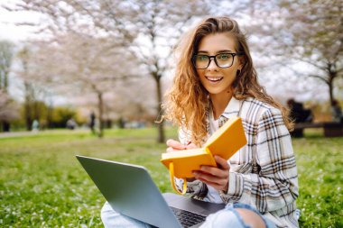 Kampüs parkındaki yeşil çayır ödevini yaparken dizüstü bilgisayar okuyan pozitif bir öğrenci. Eğitim çevrimiçi. IT, uzaktan kumanda. İş, blog yazma, serbest çalışma kavramı.