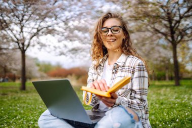 Kampüs parkındaki yeşil çayır ödevini yaparken dizüstü bilgisayar okuyan pozitif bir öğrenci. Eğitim çevrimiçi. IT, uzaktan kumanda. İş, blog yazma, serbest çalışma kavramı.
