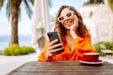 Yazlık kafede kahve içen ve cep telefonu kullanan genç bir kadın. İnsanlar, moda, yaşam tarzı, seyahat ve tatil kavramı. 