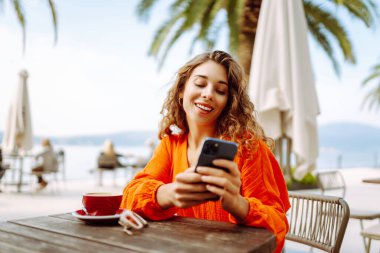 Yazlık kafede kahve içen ve cep telefonu kullanan genç bir kadın. İnsanlar, moda, yaşam tarzı, seyahat ve tatil kavramı. 