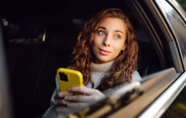 Mutlu kadın arabanın arka koltuğunda otururken akıllı telefon kullanıyor. Genç kadın arabada postaları, mesajları, blogları kontrol ediyor. İş, teknoloji kavramı.