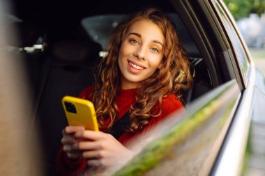 Mutlu kadın arabanın arka koltuğunda otururken akıllı telefon kullanıyor. Genç kadın arabada postaları, mesajları, blogları kontrol ediyor. İş, teknoloji kavramı.