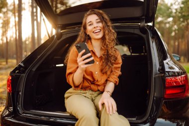 Genç bir kadın bir arabanın bagajında oturuyor ve akıllı telefon kullanıyor. Arabayla seyahat etme kavramı, aktif yaşam tarzı, blog yazma.