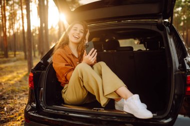 Genç bir kadın bir arabanın bagajında oturuyor ve akıllı telefon kullanıyor. Arabayla seyahat etme kavramı, aktif yaşam tarzı, blog yazma.