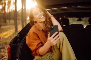 Genç bir kadın bir arabanın bagajında oturuyor ve akıllı telefon kullanıyor. Arabayla seyahat etme kavramı, aktif yaşam tarzı, blog yazma.
