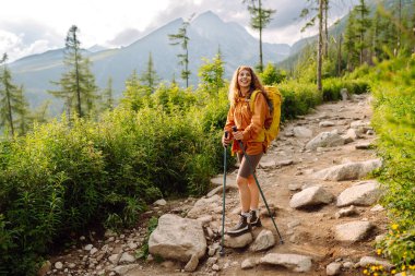 Yaz dağlarında gezintiye çıkmış bir gezgin. Sarı sırt çantalı ve yürüyüş ekipmanlı genç bir kadın dağ manzarasına karşı yürüyüş parkurunda yürüyor..