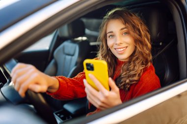 Direksiyon başında telefona bakan neşeli bir kadının portresi. Arabanın sürücüsü bir akıllı telefon kullanıyor. Boş zaman, seyahat, teknoloji, navigasyon.