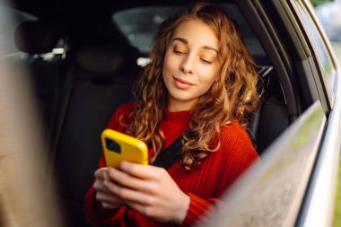 Akıllı telefon kullanan mutlu bir kadın, arabanın arka koltuğunda oturuyor, sosyal medya haberlerini sarıyorum, e-posta mektuplarını okuyorum, arkadaşlarla iletişim kuruyorum, mesajlar alıyorum, online alışveriş yapıyorum..