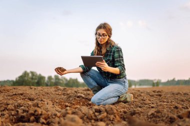 Dijital tabletli çiftçi kadın elinde kara toprak tutuyor ve kalitesini kontrol ediyor. Teknoloji, ekoloji ve bahçıvanlık kavramı.