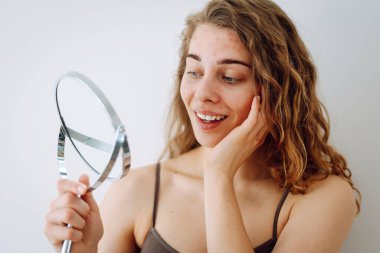 Kırmızı benekli genç bir kadın sivilceden ve deri kusurlarından mutsuz. Banyodaki aynada yansımaya bakıyor. Yüz bakımı. Sivilce sorunu.