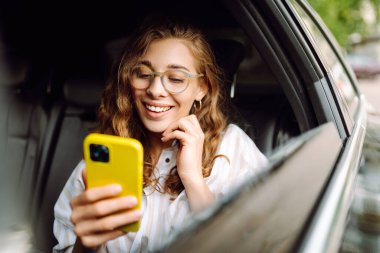 Genç bir kadın arabanın arka koltuğunda otururken akıllı telefon kullanır. Varış yerine giden yolda giderken arabada mutlu bir kadın. Teknoloji, blog yazma, iş konsepti.